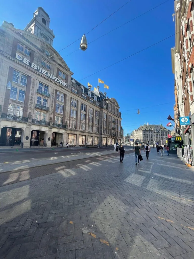 Winkelpand De Bijenkorf aan het Damrak in Amsterdam op een zonnige dag met wandelaars op straat.