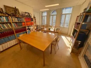 Licht studeer- of vergaderruimte aan het Damrak met houten tafels, boekenplanken en grote ramen die uitzicht bieden op de straat.