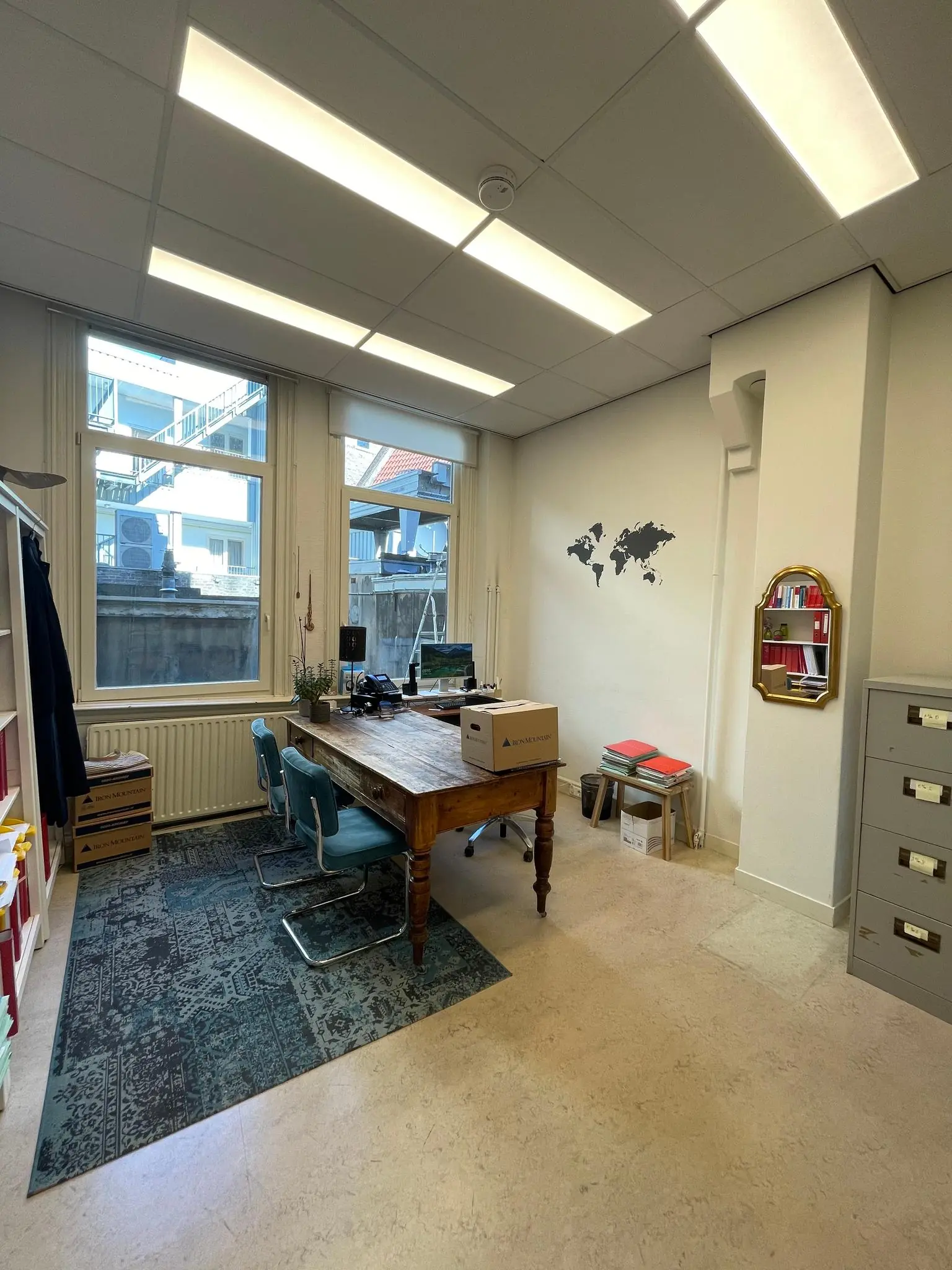 Kleine kantoorruimte aan het Damrak met een houten bureau, twee stoelen, een wereldkaart aan de muur en archiefkasten.