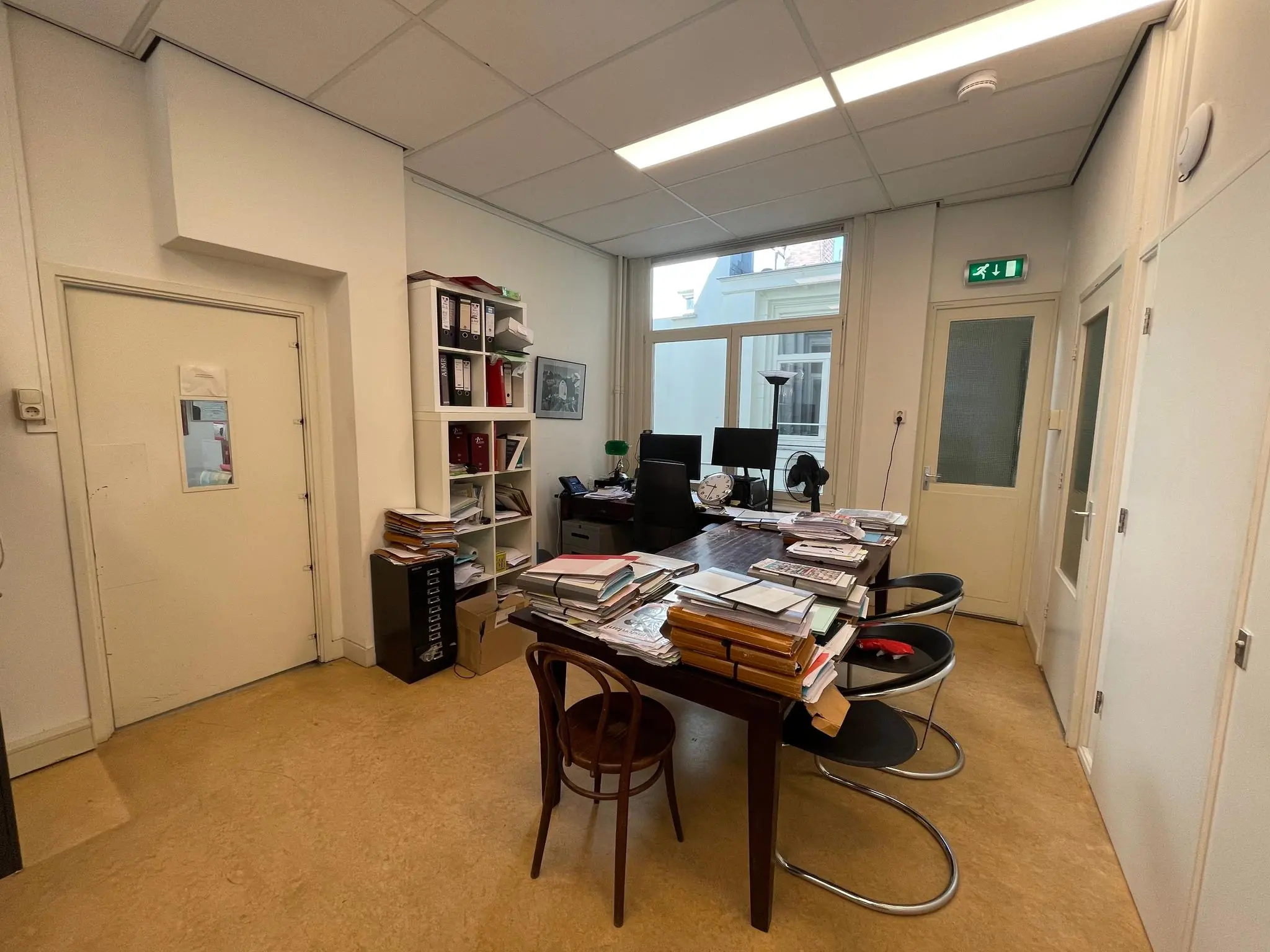 Kantoorruimte aan het Damrak met een tafel vol stapels dossiers en kantoormeubilair.