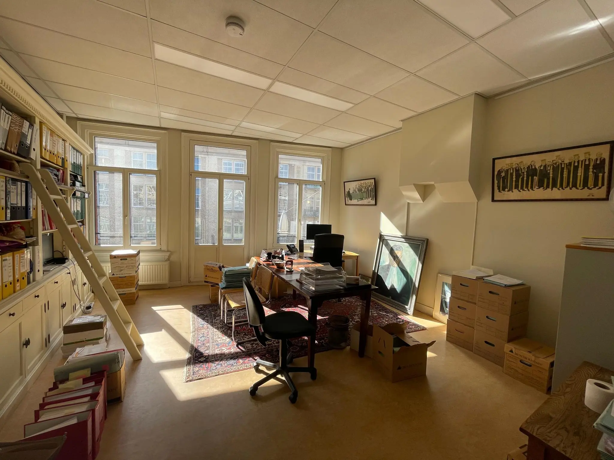 Zonnig kantoor aan het Damrak met een klassiek interieur, stapels dossiers en grote ramen met uitzicht op Amsterdamse gevels.