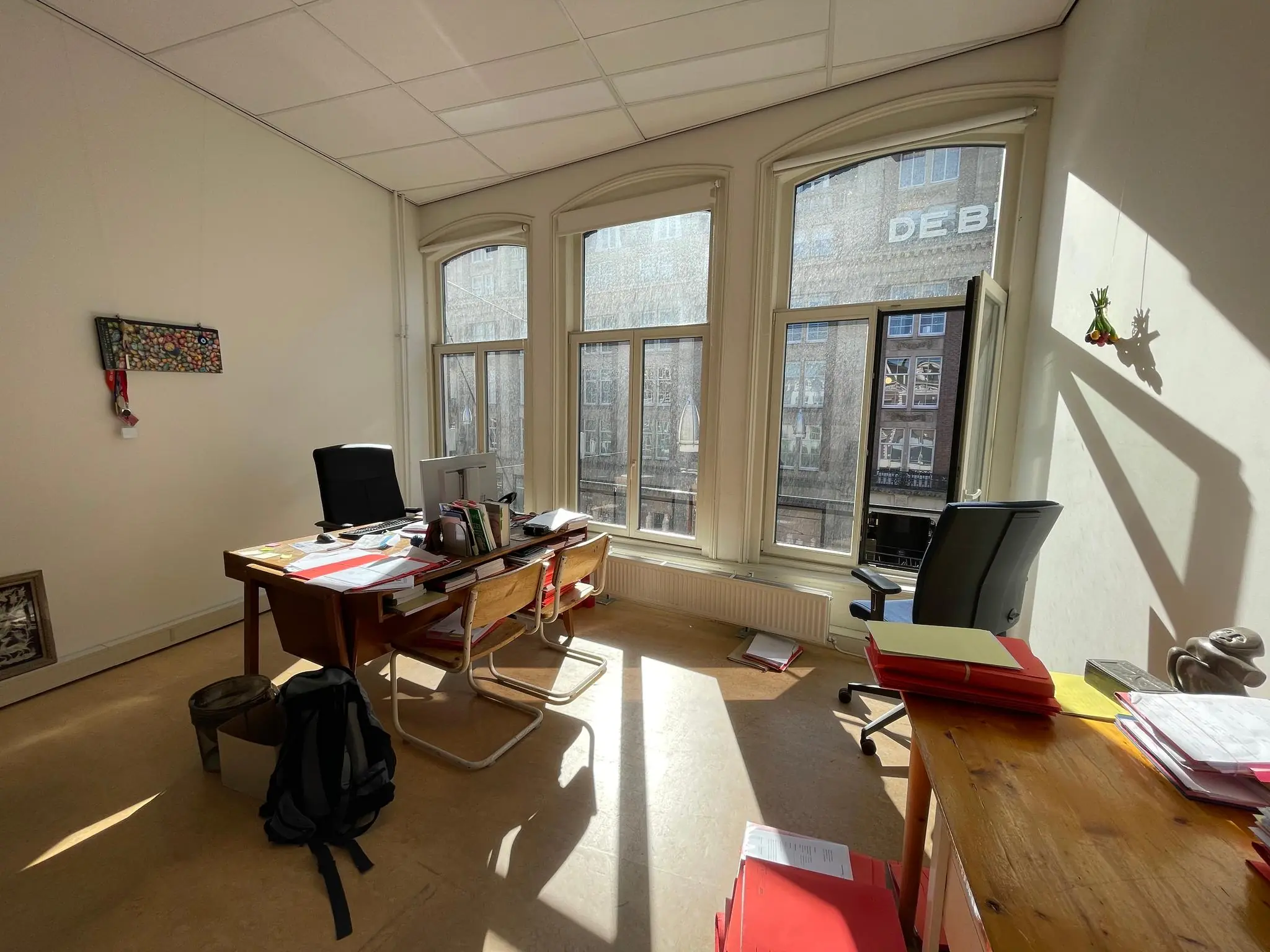Zonnig kantoor aan het Damrak met drie grote ramen, bureaus en stapels dossiers.