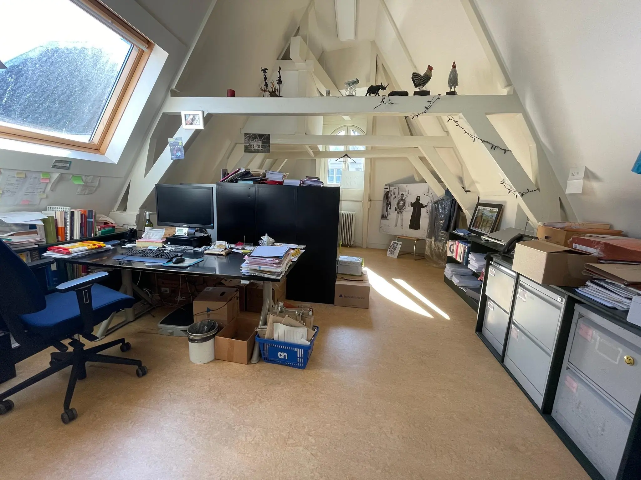 Zolderkantoor aan het Damrak met schuine dakramen, bureaus, stapels papieren en persoonlijke decoraties.
