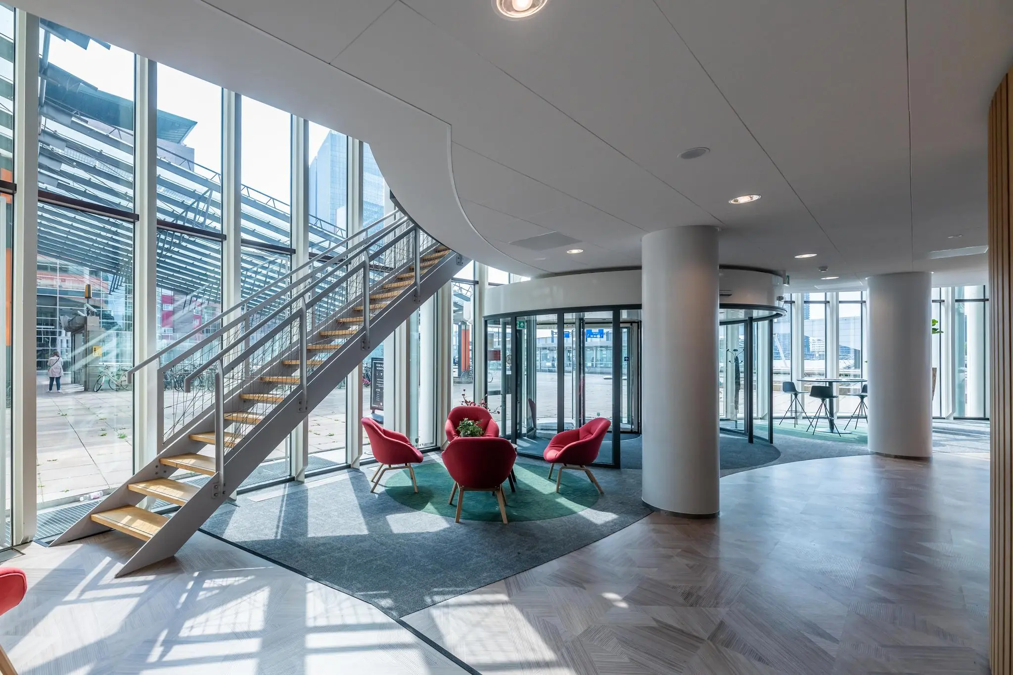 Moderne lobby met glazen gevel aan het Wilhelminaplein, voorzien van rode stoelen, een wenteltrap en een draaideur.