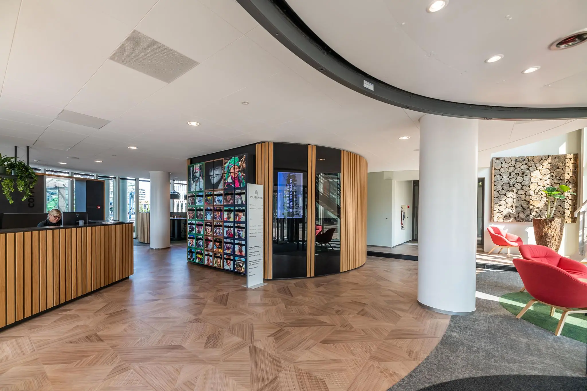 Moderne ontvangsthal van gebouw Wilhelminaplein met houten vloer, receptiebalie, kleurrijke kunst en rode zitstoelen.