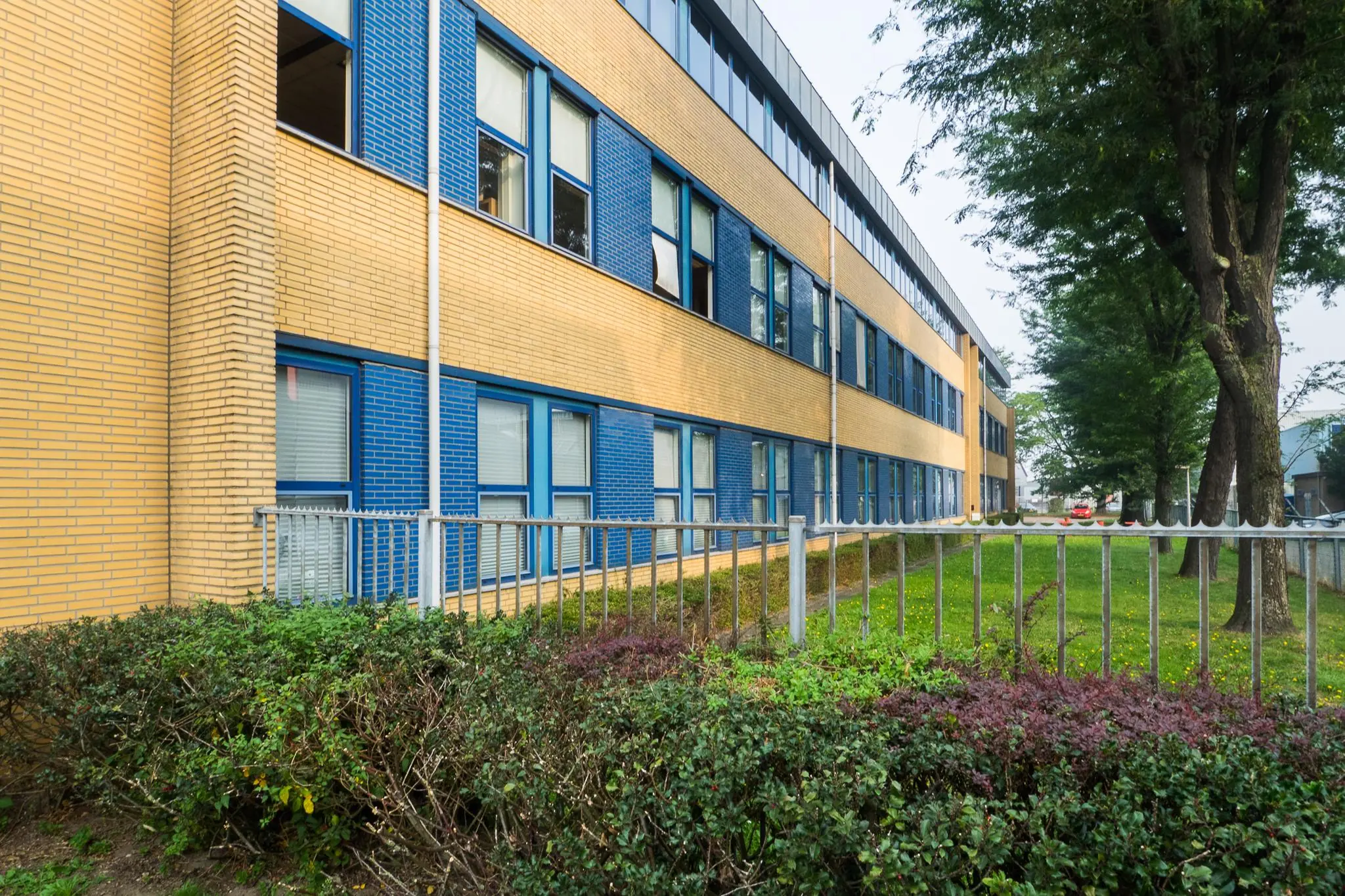 Buitenaanzicht van een modern kantoorgebouw aan de Kernkade met geel en blauw metselwerk, omringd door groen en een metalen hek.