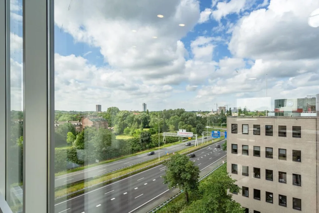 Uitzicht vanuit een kantoorgebouw aan de Euclideslaan op een snelweg, groen landschap en omliggende gebouwen onder een deels bewolkte lucht.