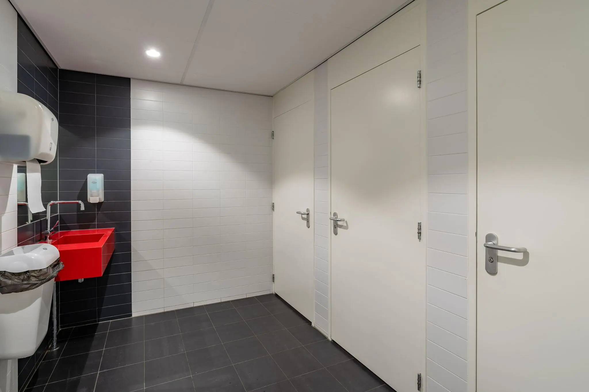 Moderne openbare toiletruimte met drie toiletdeuren, een felrood wastafelmeubel en een zwart-wit betegelde wand.