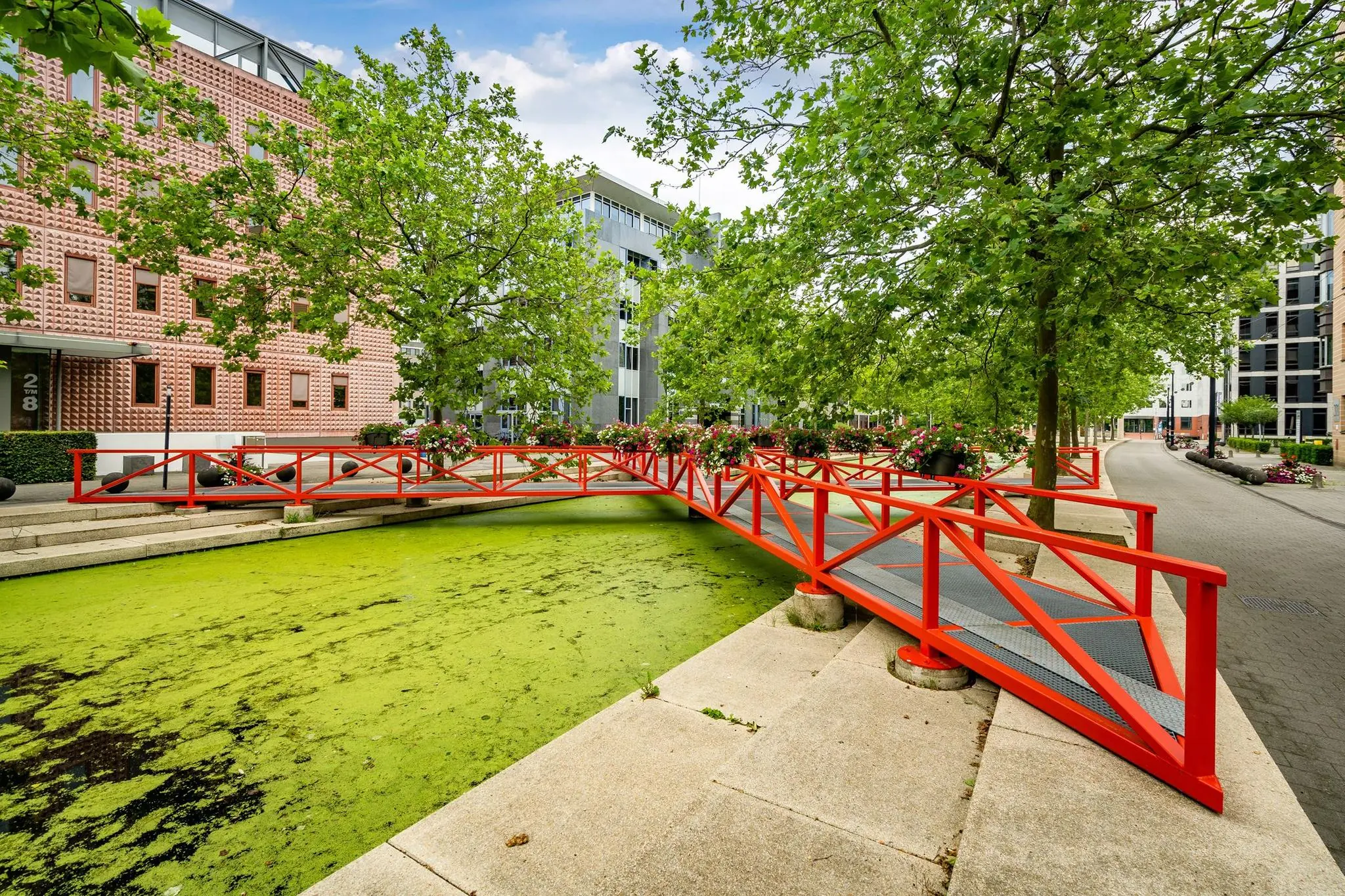 Felrode voetgangersbrug over een met algen bedekte gracht aan de Euclideslaan, omgeven door bomen en moderne kantoorgebouwen.