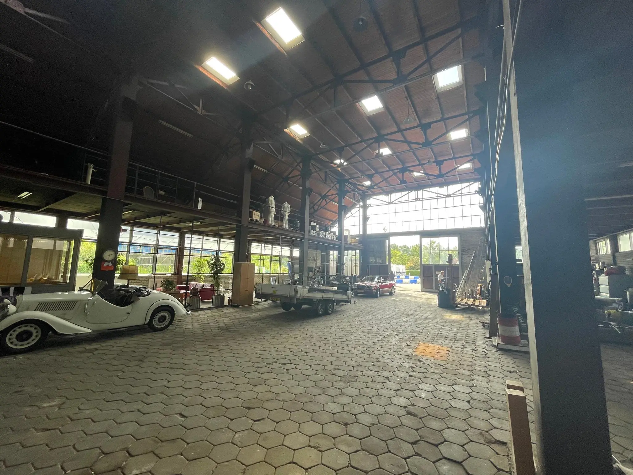Interieur van een ruime loods aan de Sloterweg met klassieke auto’s, een trailer en hoge ramen die daglicht binnenlaten.