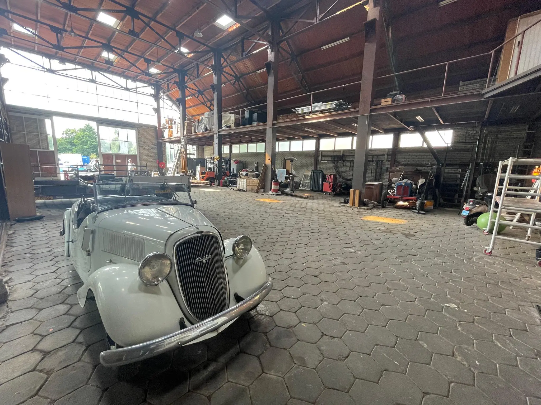 Witte oldtimer in een ruime, industriële loods aan de Sloterweg met diverse werkmaterialen en voertuigen op de achtergrond.