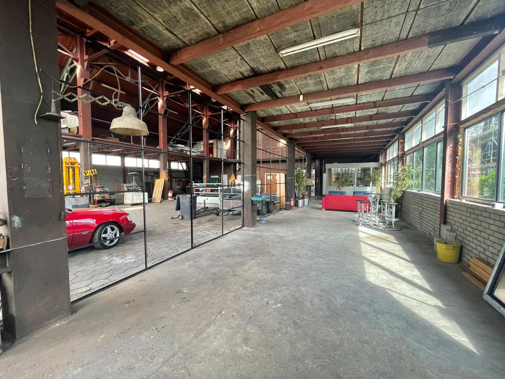 Interieur van een ruime, industriële loods aan de Sloterweg met grote ramen, een betonnen vloer en zicht op een rode auto in een aangrenzende ruimte.