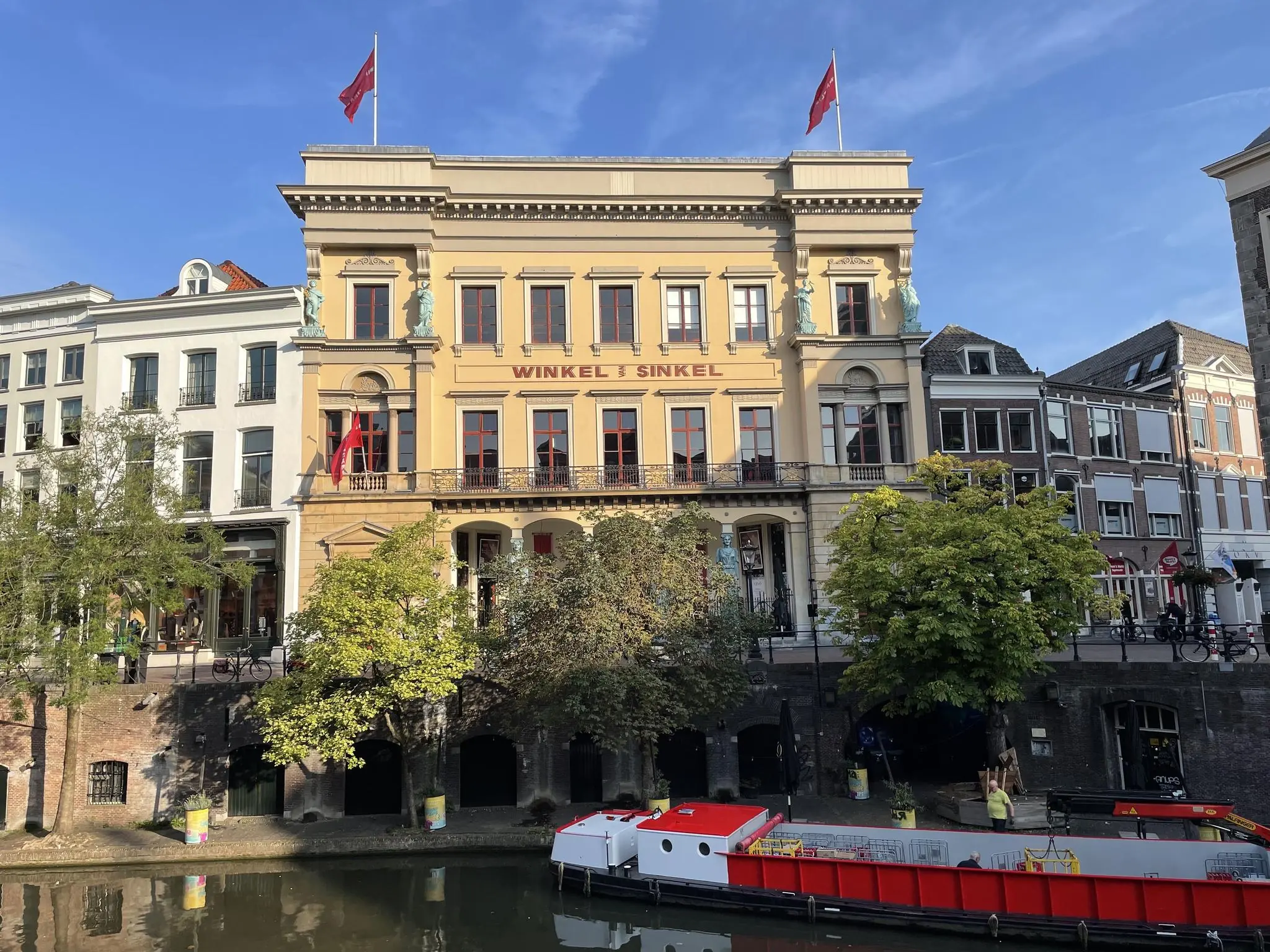 Gevel van het historische pand Winkel van Sinkel aan de Oudegracht in Utrecht, met bomen en een rood-witte rondvaartboot op de voorgrond.