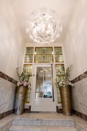 Een elegante hal met een kristallen kroonluchter, marmeren trap en twee grote vazen met bloemen naast een glazen deur met gebrandschilderd glas.