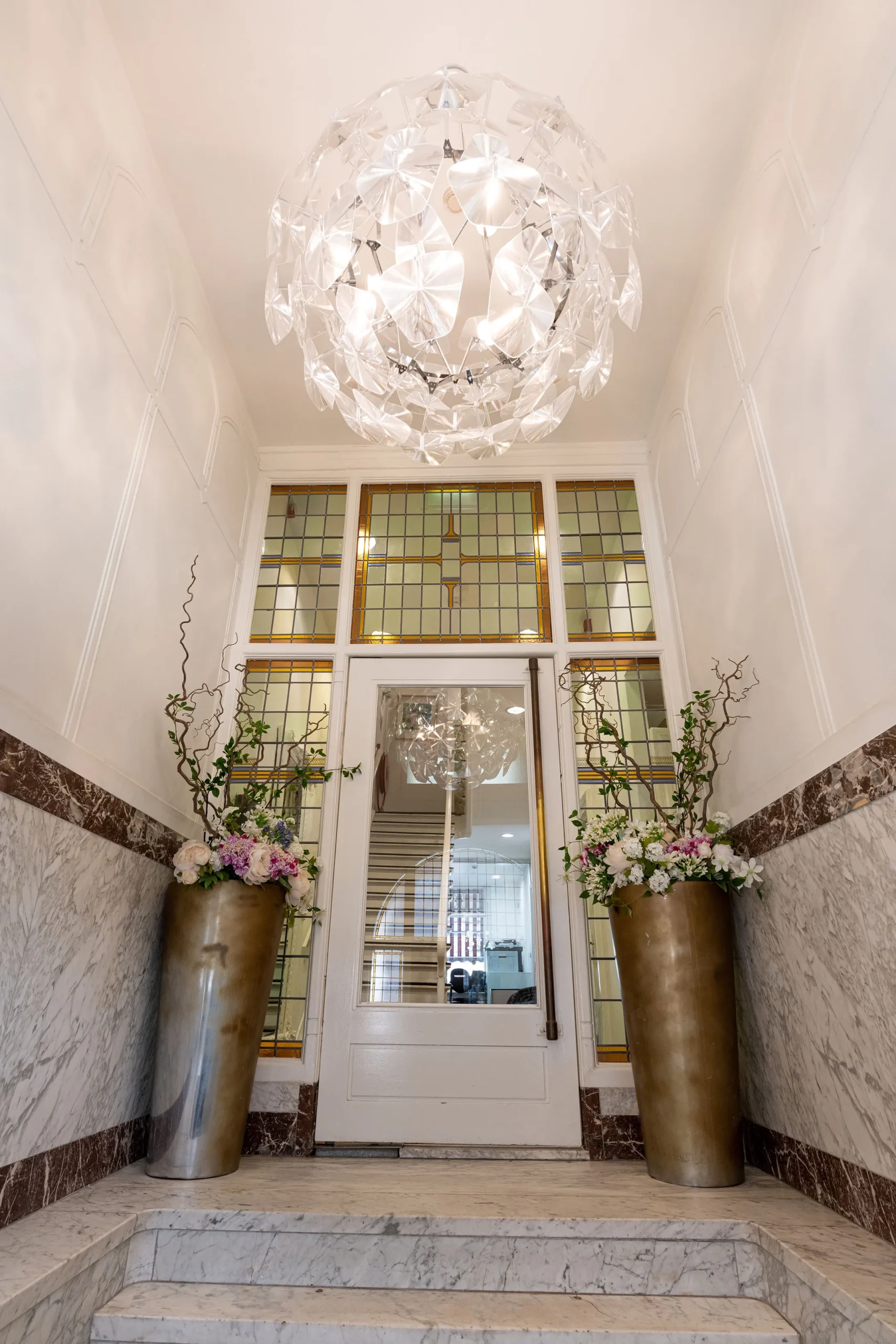 Een elegante hal met een kristallen kroonluchter, marmeren trap en twee grote vazen met bloemen naast een glazen deur met gebrandschilderd glas.