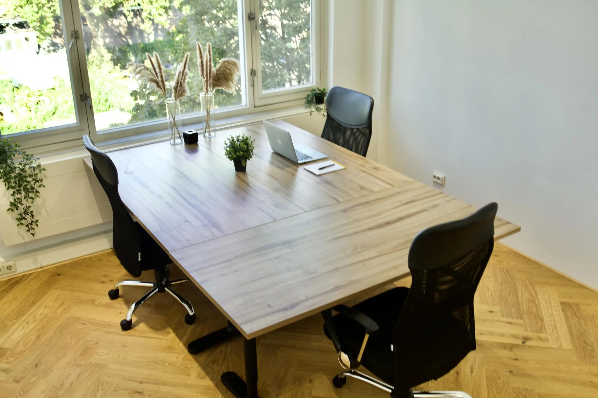 Modern kantoor met houten vergadertafel, vier zwarte stoelen en uitzicht op groene tuin via grote ramen.