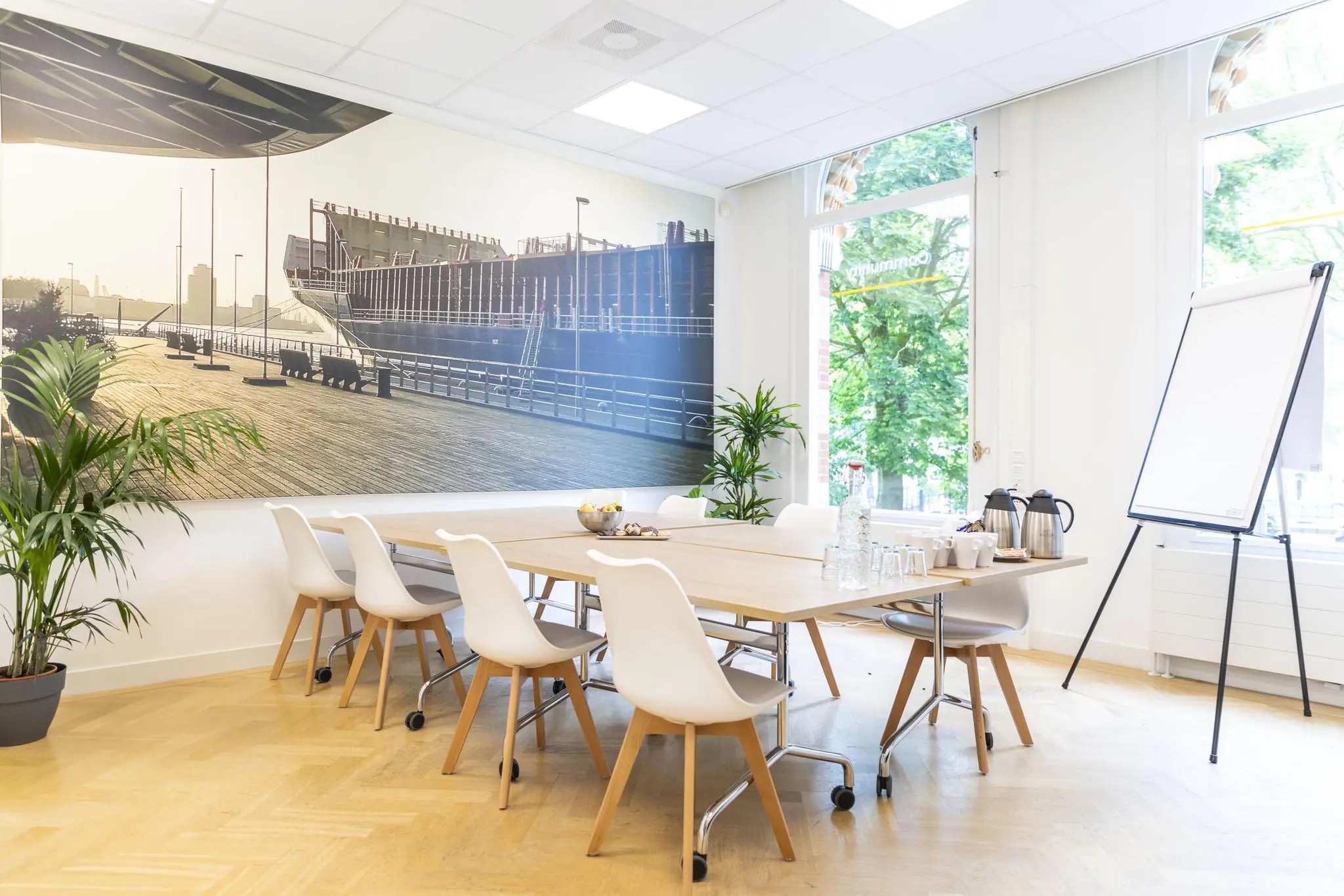 Moderne vergaderruimte met een grote wandfoto van het Westplein, witte stoelen, houten tafel en een flip-over.