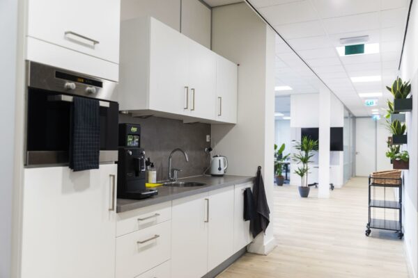 Moderne kantoorpantry met witte kasten, koffiezetapparaat en uitzicht op een lichte gang met planten.