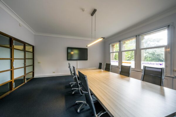 Vergaderruimte met lange houten tafel, moderne stoelen, een scherm aan de muur en ramen met uitzicht op Emmaplein.