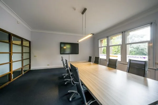 Vergaderruimte met lange houten tafel, moderne stoelen, een scherm aan de muur en ramen met uitzicht op Emmaplein.