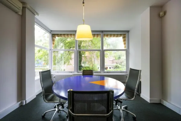 Vergaderruimte aan het Emmaplein met een ronde blauw-oranje tafel, vier stoelen en grote ramen met uitzicht op groen.