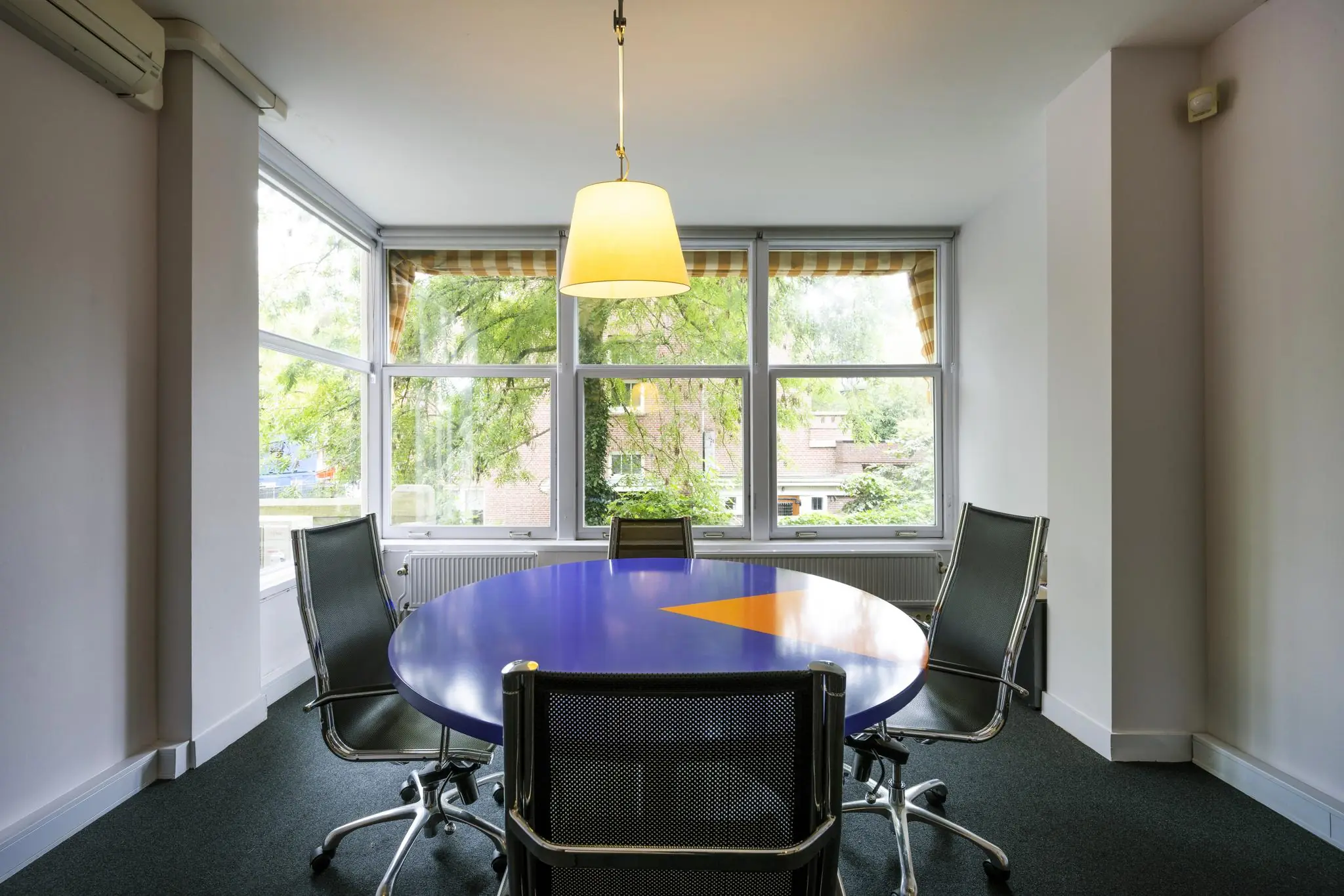 Vergaderruimte aan het Emmaplein met een ronde blauw-oranje tafel, vier stoelen en grote ramen met uitzicht op groen.