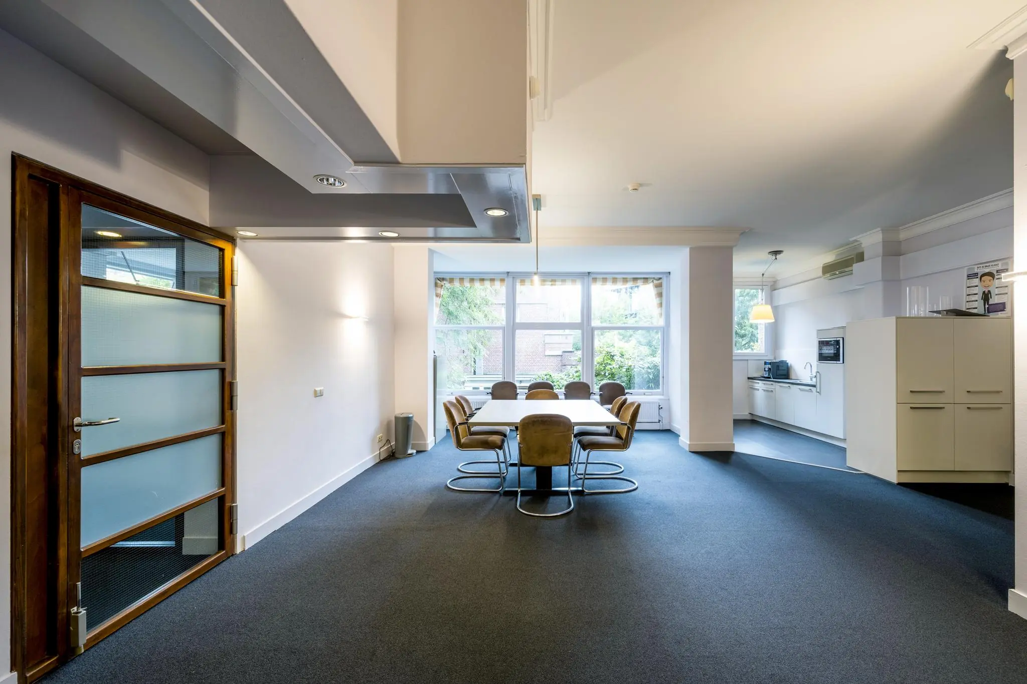 Vergaderruimte aan het Emmaplein met een grote tafel, acht stoelen, een keukenblok en grote ramen met uitzicht op groen.