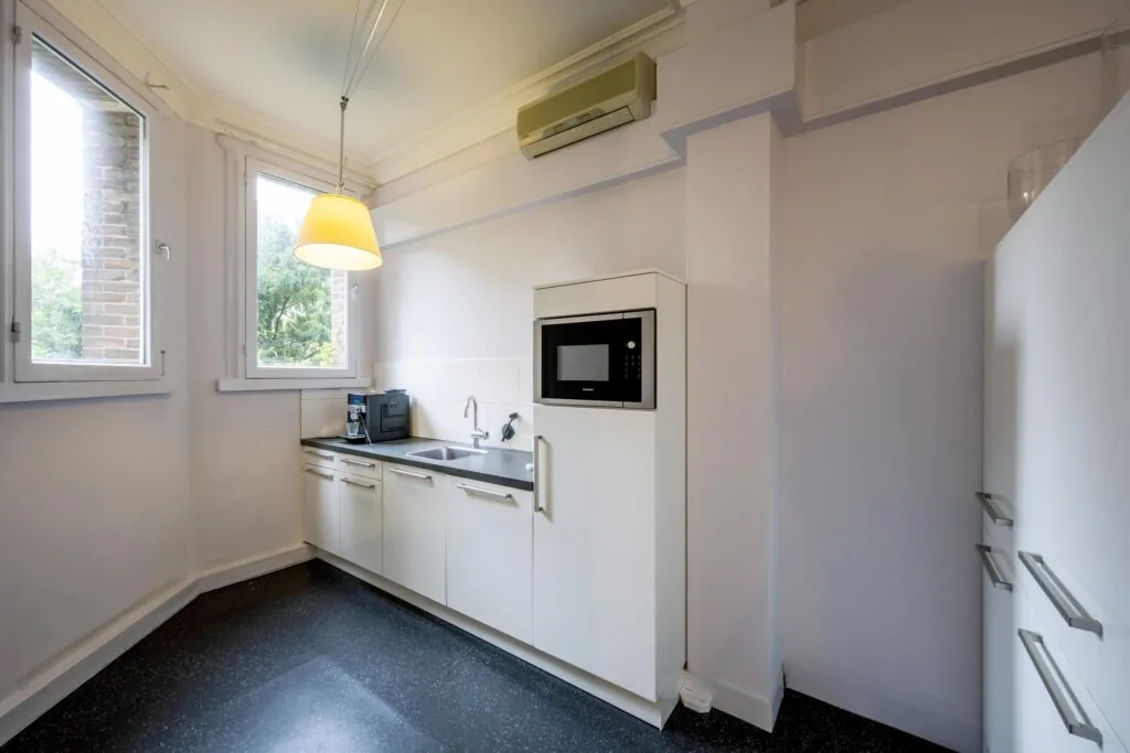 Moderne witte keuken met ingebouwde magnetron, koffiezetapparaat en twee ramen die uitzicht bieden op groen.