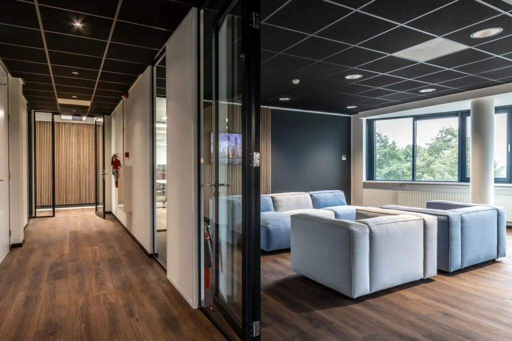 Moderne kantoorruimte met een zithoek met lichtblauwe fauteuils en grote ramen die uitzicht bieden op groen.