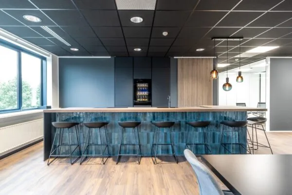 Moderne kantoorkeuken met een lange bar, zwarte barkrukken, hanglampen en een ingebouwde koelkast.