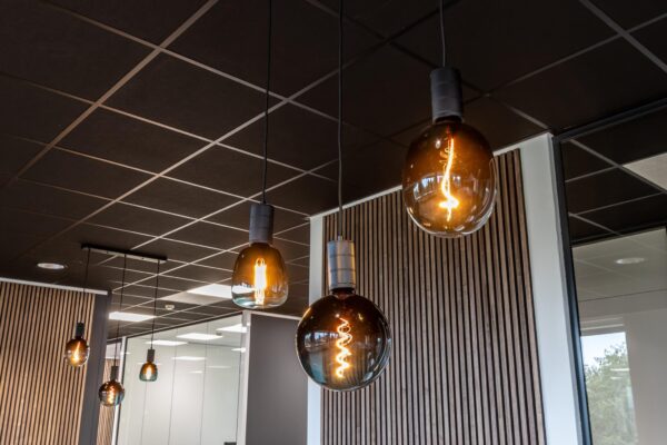 Moderne hanglampen met warm licht hangen aan een zwart systeemplafond in een strak ingericht kantoor.