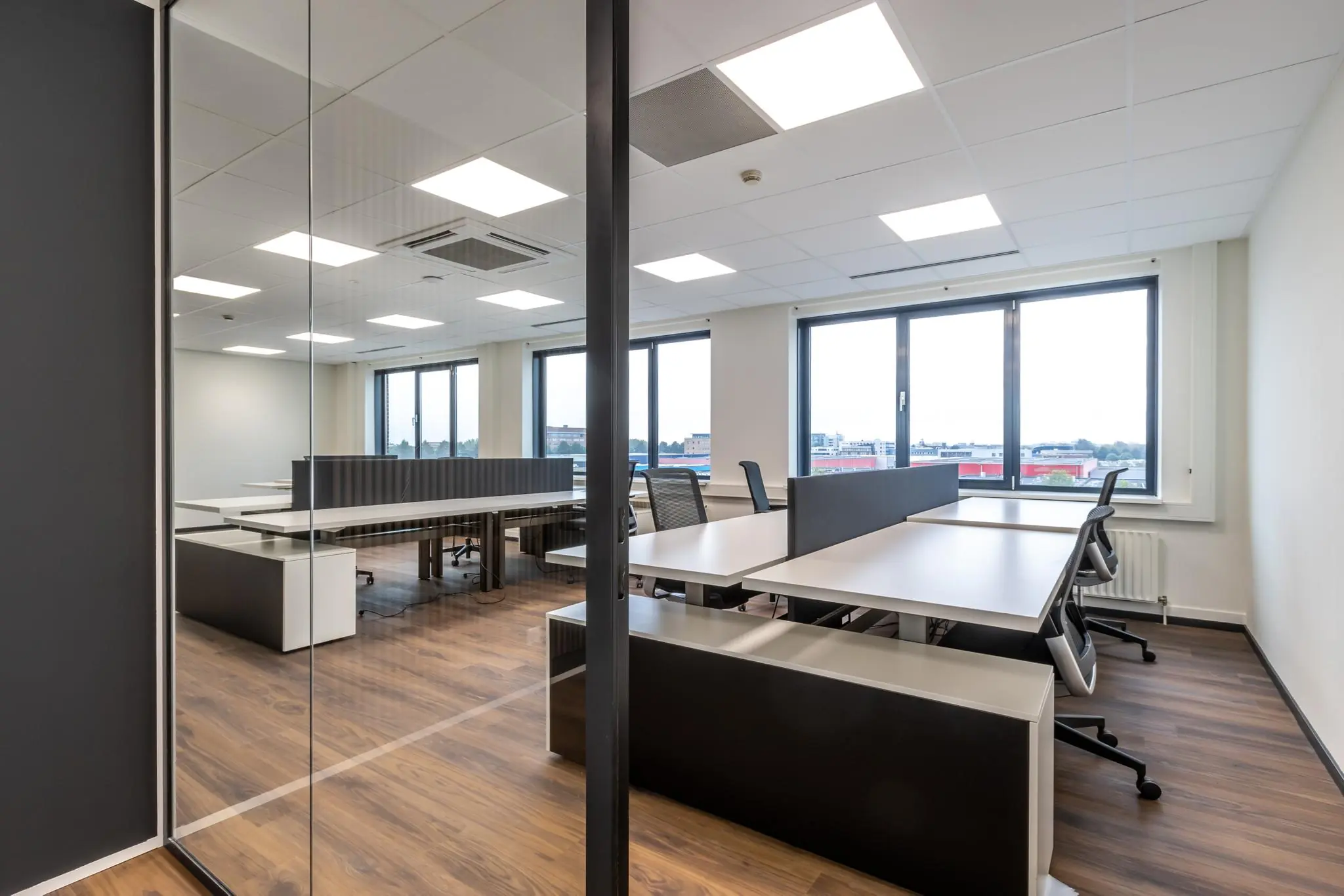 Modern kantoor met open werkplekken, bureaustoelen en grote ramen met uitzicht op een bedrijventerrein.
