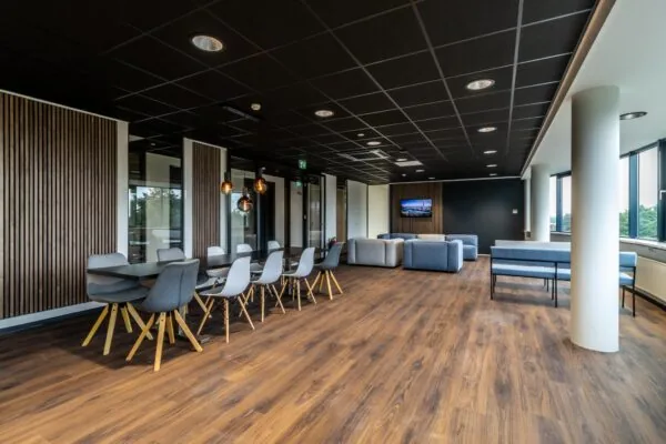 Moderne kantoorlounge met houten vloer, designmeubilair en grote ramen die uitzicht bieden op groen.