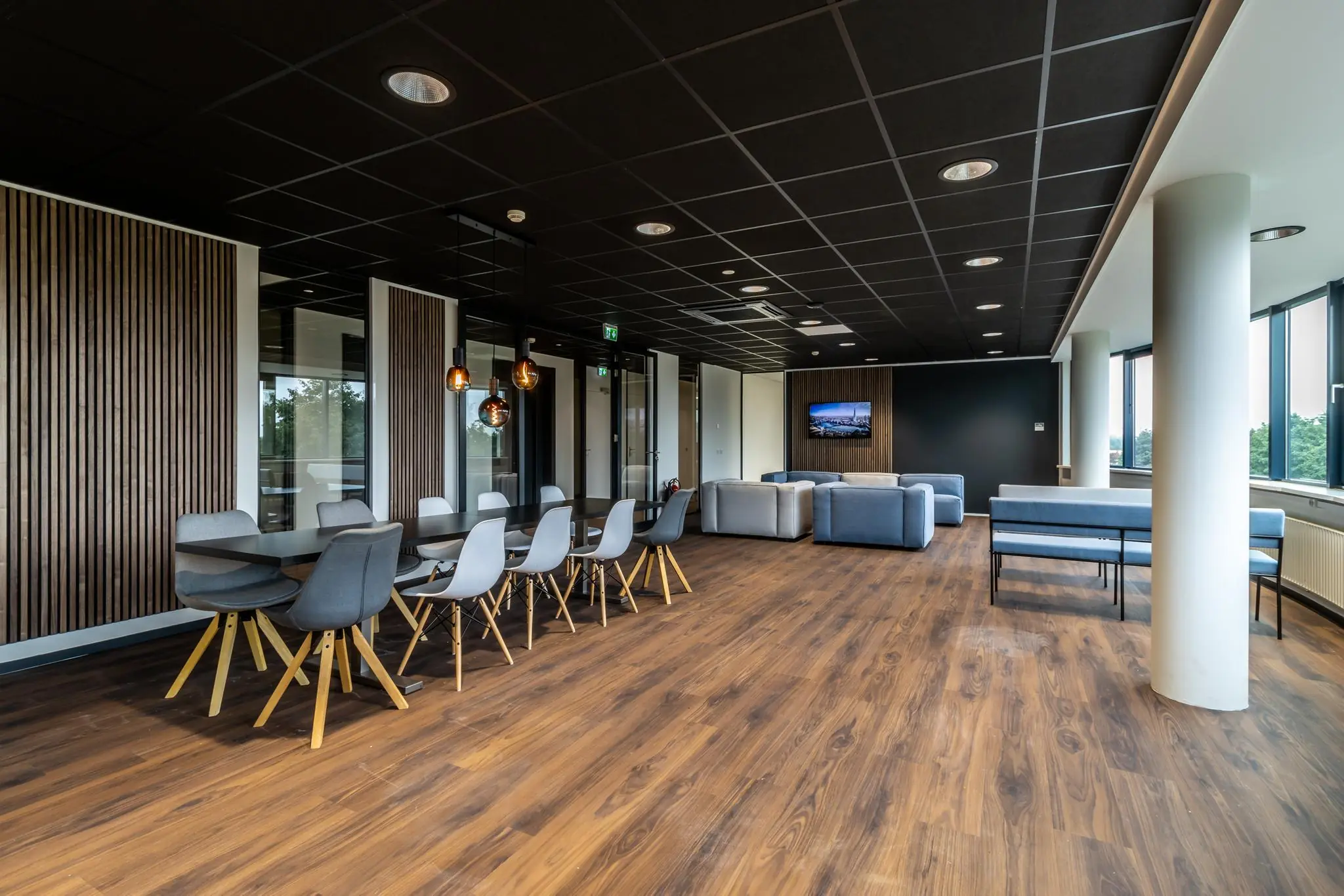 Moderne kantoorlounge met houten vloer, designmeubilair en grote ramen die uitzicht bieden op groen.