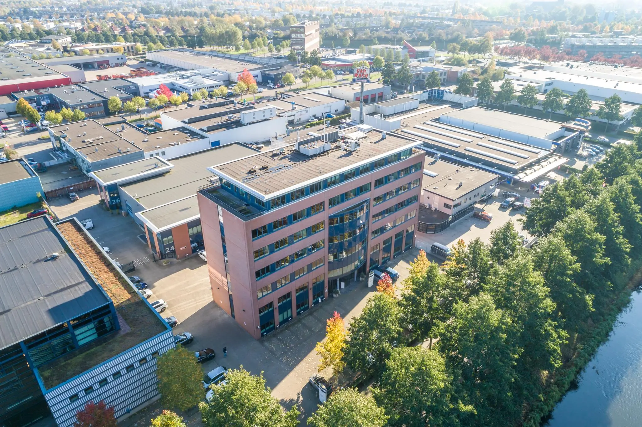 Luchtfoto van een rood kantoorgebouw in een bedrijventerrein met omliggende bedrijfsgebouwen en bomen langs een waterkant.