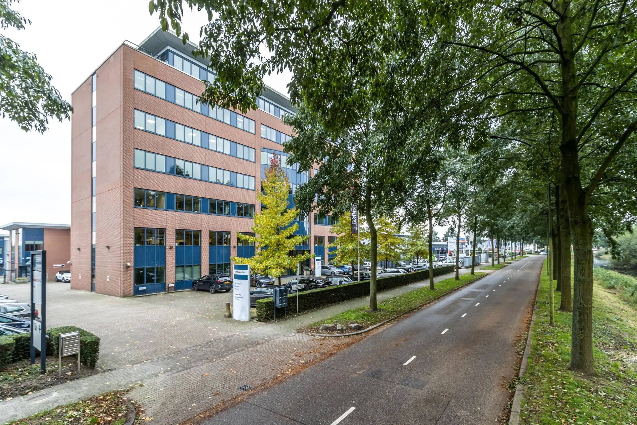 Kantoorgebouw aan een boomrijke straat met parkeergelegenheid en bewegwijzering.