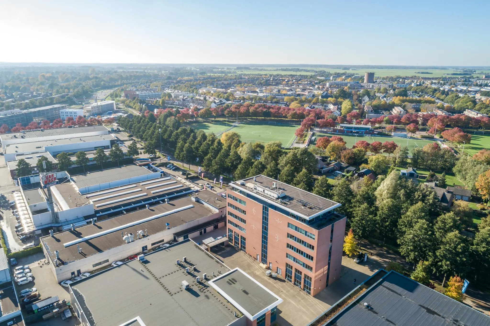 Luchtfoto van een bedrijventerrein met een rood bakstenen kantoorgebouw en een naastgelegen sportveld omringd door herfstbomen.