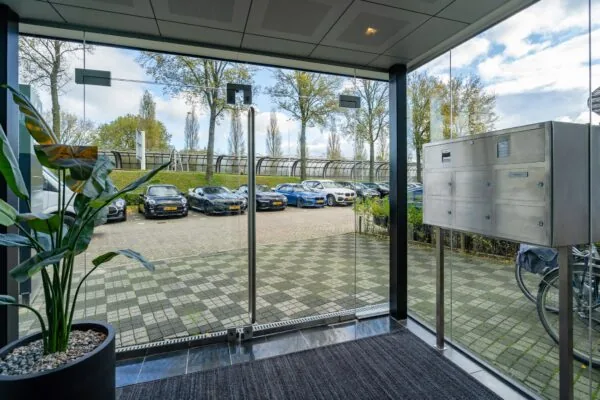 Ingang van een modern kantoorpand aan de Mijlweg met uitzicht op een parkeerterrein vol geparkeerde auto’s.