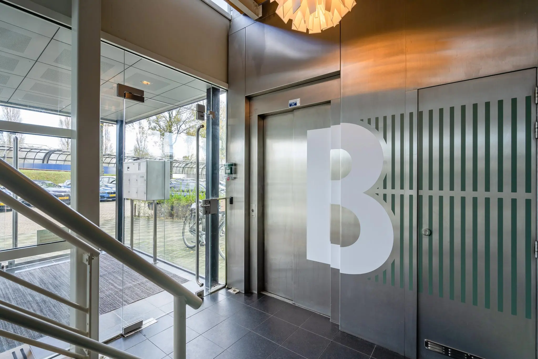 Ingang van een modern kantoorgebouw aan de Mijlweg met een lift, glazen deuren en de letter B op een metalen wand.