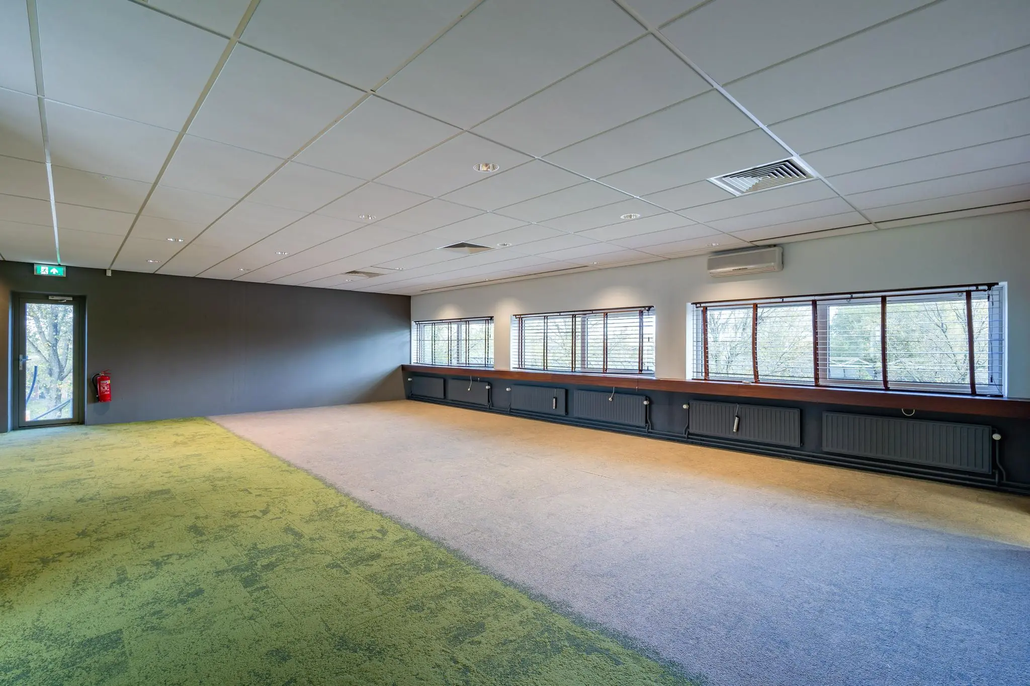 Lege kantoorruimte aan de Mijlweg met tapijtvloer, systeemplafond en grote ramen met uitzicht op bomen.