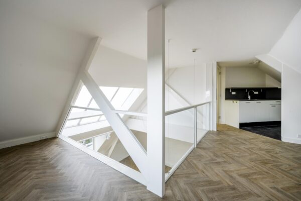 Zolderverdieping van een modern appartement aan de Emmalaan met visgraatvloer, een glazen balustrade en een open keuken.