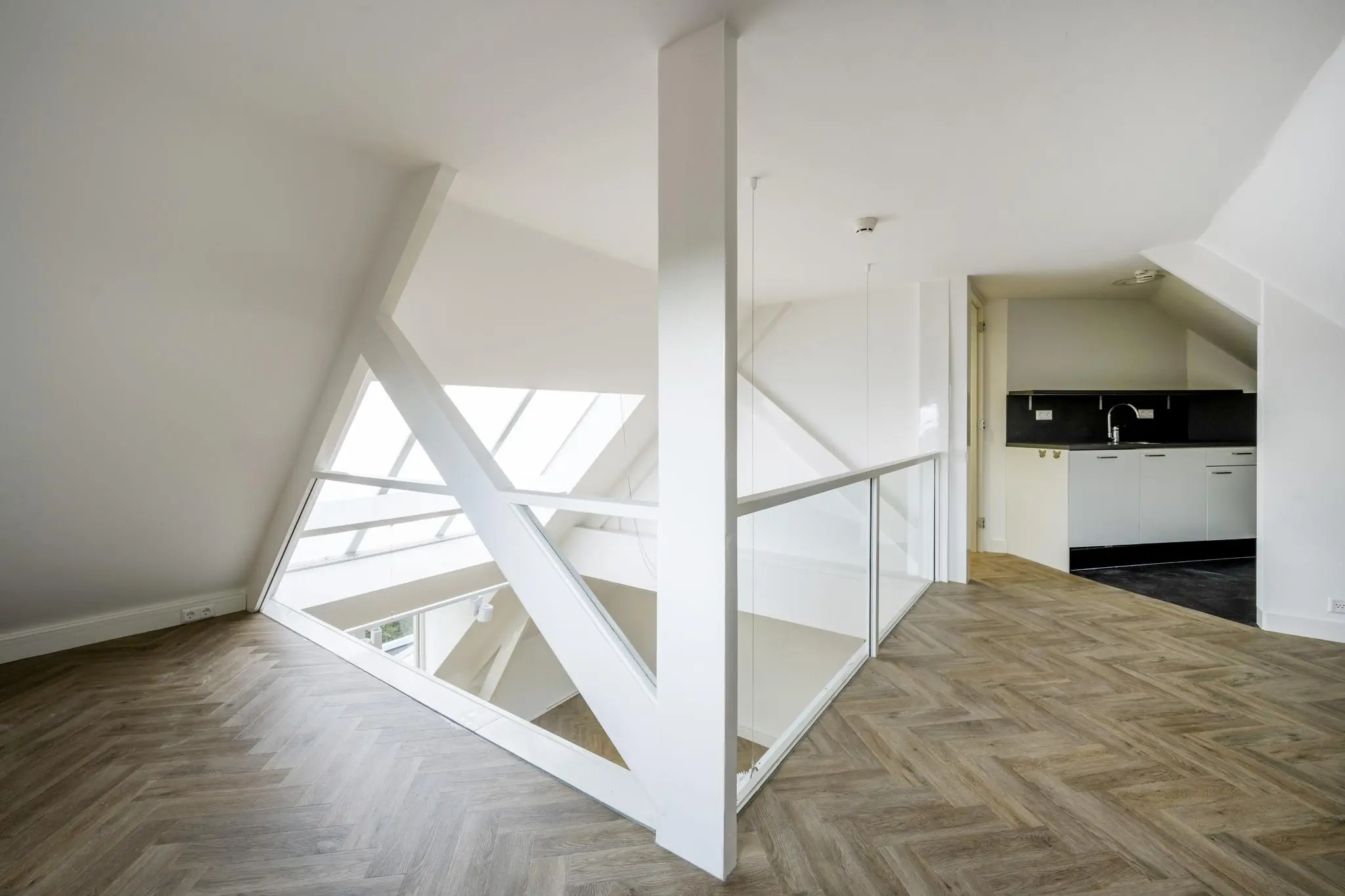Zolderverdieping van een modern appartement aan de Emmalaan met visgraatvloer, een glazen balustrade en een open keuken.