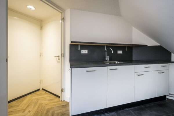 Moderne kitchenette met witte kasten, zwarte blad en aangrenzende hal met houten visgraatvloer.