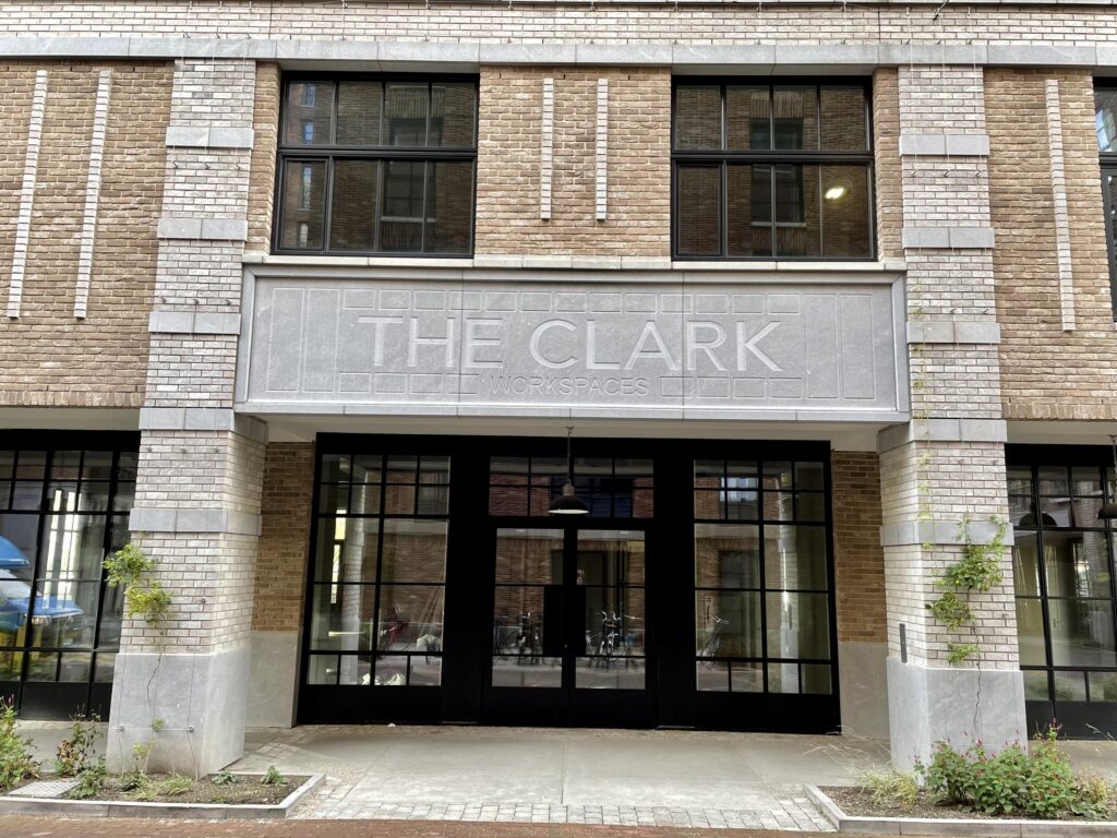 Een bakstenen gebouw met grote ramen en een bord met de tekst "THE CLARK WORKSPACES" boven de ingang.