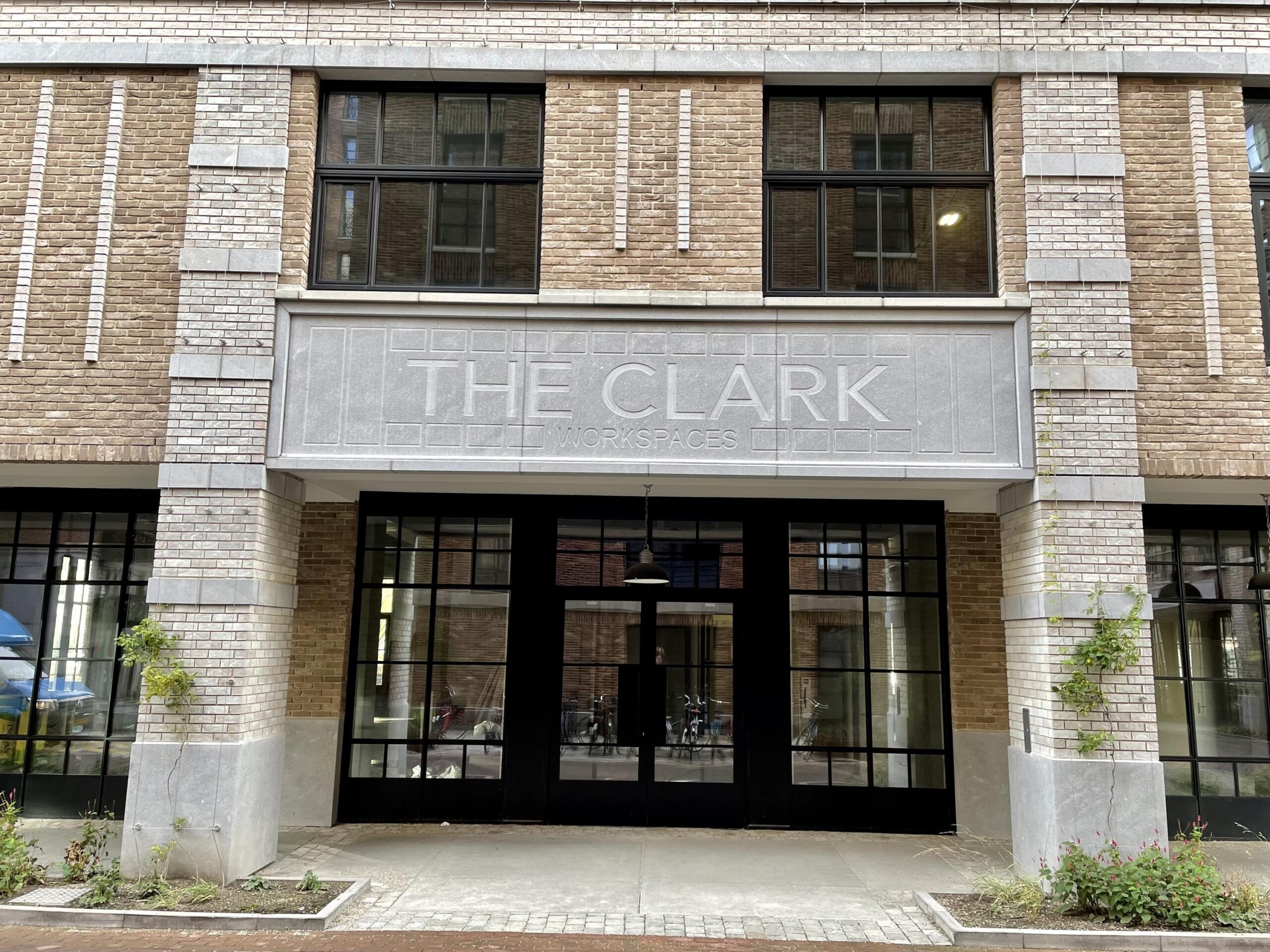 Een bakstenen gebouw met grote ramen en een bord met de tekst "THE CLARK WORKSPACES" boven de ingang.