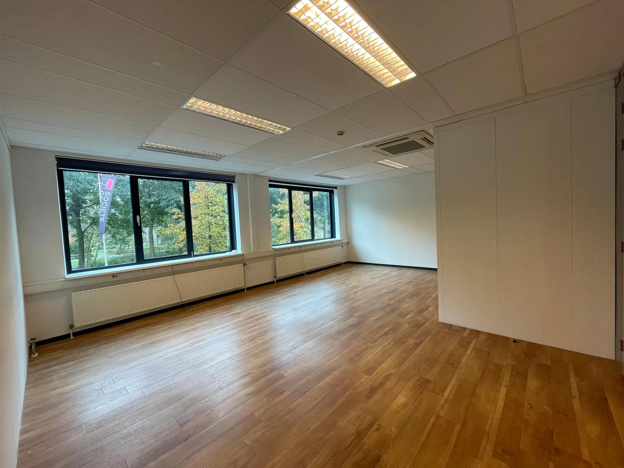 Licht en leeg kantoor met houten vloer, grote ramen en een neutraal interieur.