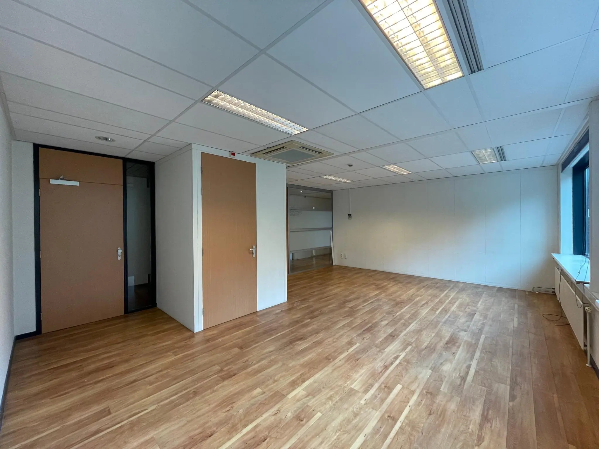 Lege kantoorruimte met houten vloer, systeemplafond en twee gesloten deuren.
