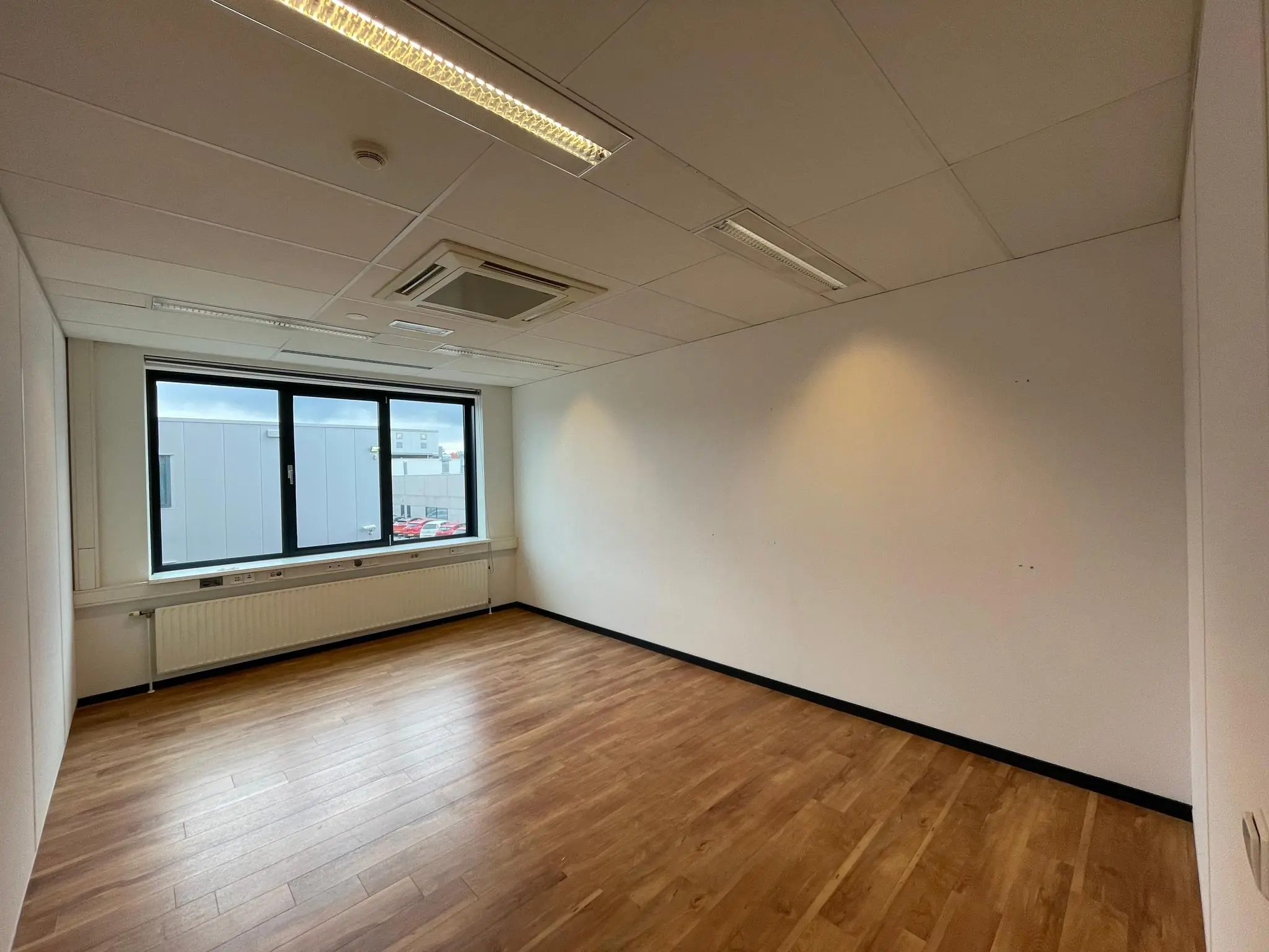 Lege kantoorruimte met houten vloer, witte muren en een groot raam met uitzicht op een industrieel gebouw.