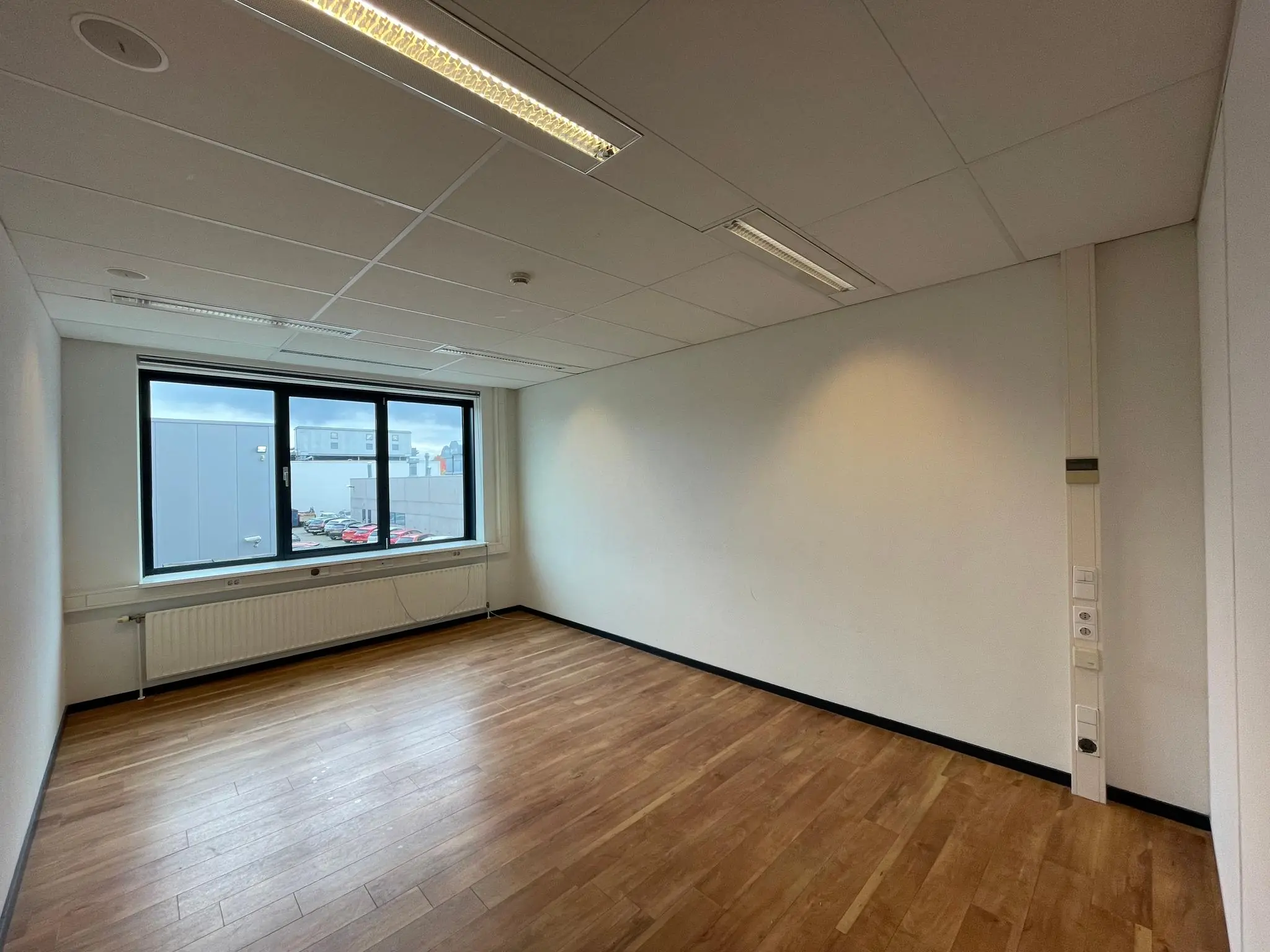 Lege kantoorruimte met houten vloer, wit plafond en een groot raam met uitzicht op een bedrijventerrein.