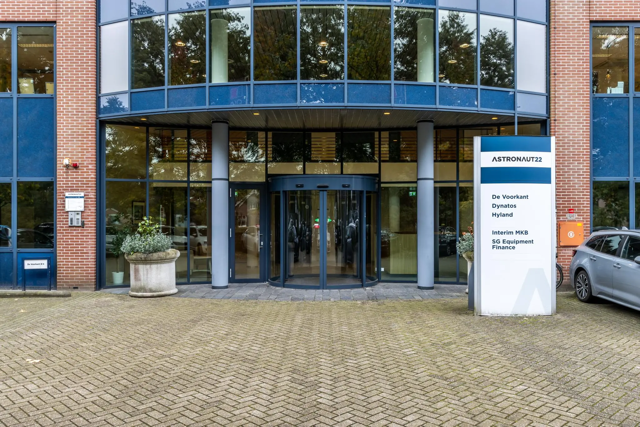Ingang van kantoorgebouw Astronaut 22 met een draaideur, bedrijfsnaambord en geparkeerde auto.
